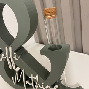 May include: A sage green ampersand symbol stand featuring white cursive text that reads "Leffi & Mathing" on the front. The structure includes a cylindrical glass test tube with a cork stopper inserted into the top of the ampersand shape.