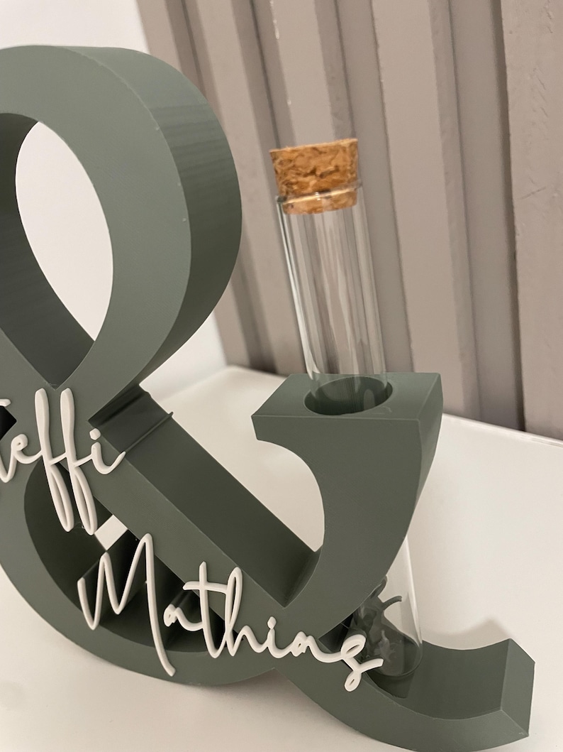 May include: A sage green ampersand symbol stand featuring white cursive text that reads "Leffi & Mathing" on the front. The structure includes a cylindrical glass test tube with a cork stopper inserted into the top of the ampersand shape.