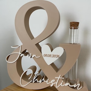 May include: A beige ampersand-shaped wooden sign featuring the white script names Jana and Christian, a white heart with the date 08.08.2026, and a clear glass test tube with a cork stopper integrated into the right side of the structure.