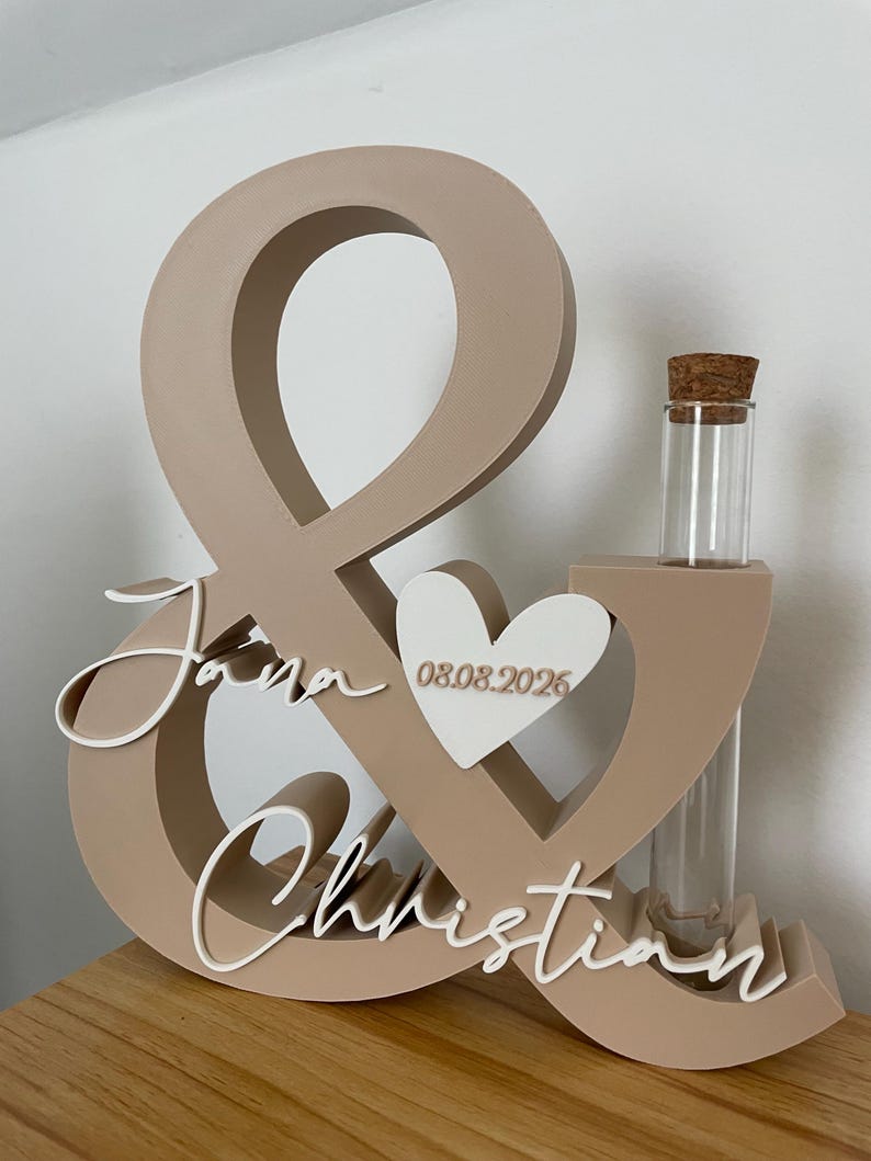 May include: A beige ampersand-shaped wooden sign featuring the white script names Jana and Christian, a white heart with the date 08.08.2026, and a clear glass test tube with a cork stopper integrated into the right side of the structure.
