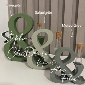 May include: Three ampersand-shaped wooden stands in olive green, sage green, and muted green. Each stand features a clear glass test tube with a cork stopper and white script text reading Stephan, Denise, and Alex Felix respectively.