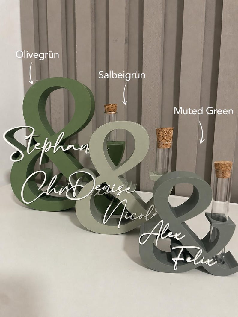 May include: Three ampersand-shaped wooden stands in olive green, sage green, and muted green. Each stand features a clear glass test tube with a cork stopper and white script text reading Stephan, Denise, and Alex Felix respectively.