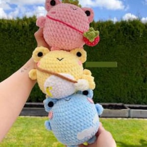 Puede incluir: Una pila de tres peluches de rana de ganchillo en rosa, amarillo y azul. La rana rosa tiene un accesorio de fresa, la rana amarilla tiene un pequeño sombrero y la rana azul tiene un detalle de nube. Las ranas están sostenidas en las manos de una persona.