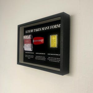 Può includere: Un espositore incorniciato con la scritta "LUXURY TAKES MANY FORMS". L'espositore include una pila di denaro, una chiave Ferrari rossa e una barra d'oro. La cornice è nera e montata su un muro bianco.