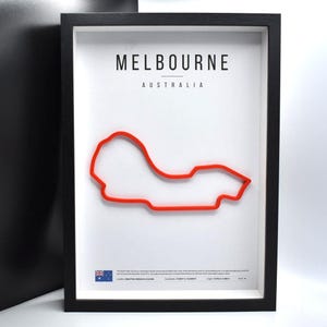 Puede incluir: Obra de arte enmarcada en negro con el texto "MELBOURNE AUSTRALIA" sobre un contorno rojo de una pista de carreras. La obra de arte está sobre un fondo blanco. La parte inferior de la obra tiene texto y una bandera.