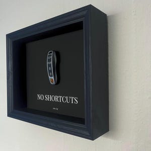 Könnte beinhalten: Gerahmtes Kunstwerk mit einem Autoschlüssel auf schwarzem Hintergrund. Der Schlüssel hat silberne und orangefarbene Akzente. Der Satz "NO SHORTCUTS" ist darunter in Weiß gedruckt. Der Rahmen ist dunkelblau.