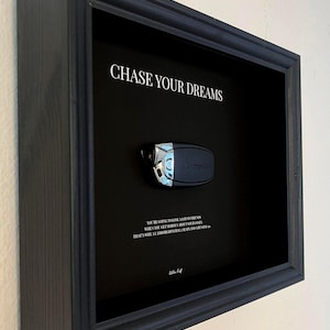 Könnte beinhalten: Gerahmtes Kunstwerk mit schwarzem Hintergrund und Autoschlüssel. Der Text "CHASE YOUR DREAMS" steht oben. Unter dem Schlüssel befindet sich zusätzlicher Text. Der Rahmen ist aus dunklem Holz.