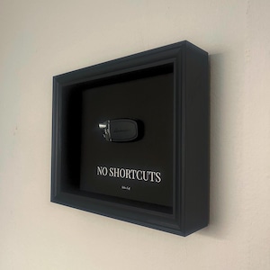 Könnte beinhalten: Ein schwarzer Schattenrahmen mit einem Autoschlüssel und den weißen Worten „NO SHORTCUTS“. Der Schlüssel ist silber und schwarz, vor einem schwarzen Hintergrund. Der rechteckige Rahmen ist an einer weißen Wand befestigt.