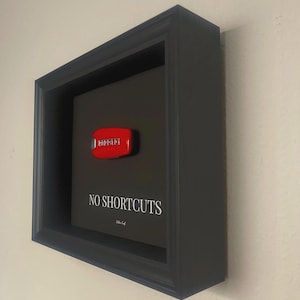Könnte beinhalten: Ein gerahmtes Kunstwerk mit einem schwarzen Rahmen und einem schwarzen Hintergrund. Das Kunstwerk zeigt den Text "NO SHORTCUTS" in Weiß, mit einem roten Ferrari-Emblem. Der Rahmen ist an einer hellen Wand befestigt.