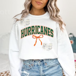 Puede incluir: Sudadera blanca con la palabra "HURRICANES" en letras verdes y naranjas. Un lazo naranja está centrado debajo de la palabra. La sudadera es de cuello redondo.