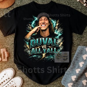May include: Black t-shirt with a graphic of a smiling person and lightning bolts. The text "DUVAL vs ALL Y'ALL" is in teal and yellow. The shirt is from "Shotts Shirts".