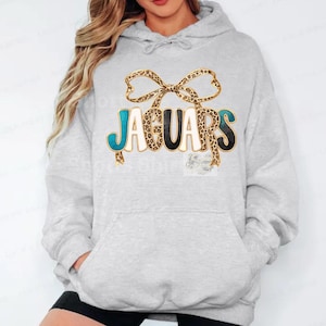 May include: Light gray hoodie with a front graphic that says "JAGUARS". The word "JAGUARS" is in teal, black, and white with a leopard print bow above the text. The hoodie has a drawstring hood and a front pocket.