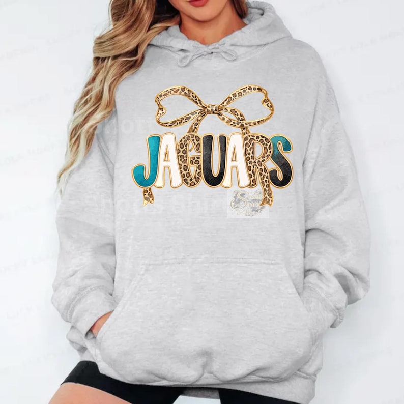 May include: Light gray hoodie with a front graphic that says "JAGUARS". The word "JAGUARS" is in teal, black, and white with a leopard print bow above the text. The hoodie has a drawstring hood and a front pocket.