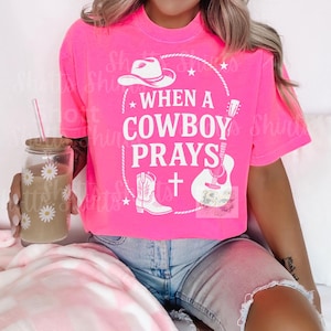 May include: Bright pink t-shirt with the text "WHEN A COWBOY PRAYS" in white, along with a cowboy hat, boots, guitar, and cross graphic. A daisy-decorated glass with a straw is held in the left hand.