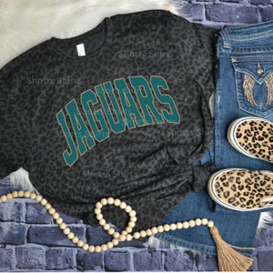 May include: A dark grey leopard print t-shirt with the word "JAGUARS" in teal and gold lettering. The shirt is styled with blue jeans, leopard print trainers, and a beaded necklace.