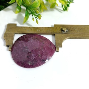 May include: A faceted, teardrop-shaped, deep red gemstone is measured with a brass caliper. The gemstone has dark inclusions and a polished surface. Green foliage is visible in the background. The caliper measures approximately 1.5 inches in length.