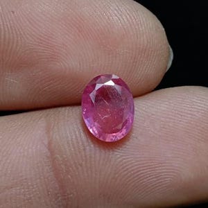 Op de afbeelding: Een roze, ovaalvormige edelsteen met facetten. De edelsteen heeft een briljante slijpvorm met meerdere facetten die het licht reflecteren. De kleur van de steen is levendig roze, met een lichte variatie in tint. De edelsteen wordt tussen twee vingers gehouden.