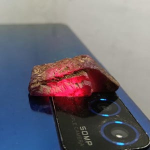 May include: A rough-cut, reddish-brown gemstone with a vibrant red interior. The stone has a textured surface and irregular shape, with a glowing red light emanating from within. The gemstone is resting on a blue surface.