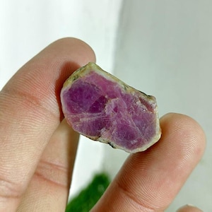 May include: A rough-cut, natural ruby gemstone. The ruby has a deep magenta color with a slightly rough texture and a light-colored outer edge. The gemstone is held between two fingers.