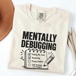 May include: A cream-colored t-shirt featuring the black text MENTALLY DEBUGGING above a checklist graphic. The list includes Finding the issue, Analyzing, Fixing (maybe), and Still thinking, accompanied by a pencil illustration next to the list.
