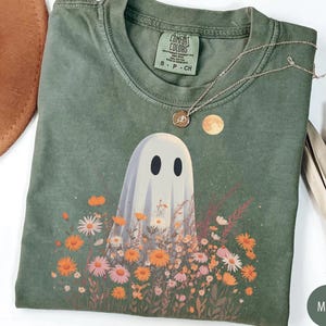 May include: A moss green t-shirt with a ghost graphic surrounded by flowers. The ghost has two black oval eyes. A gold necklace with a pendant and a small moon graphic are on the shirt. The shirt is folded.