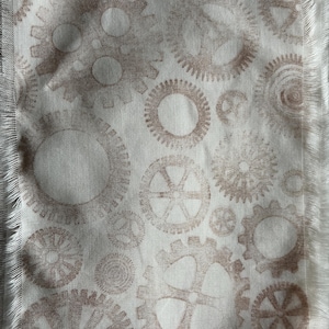 May include: Cream-coloured fabric featuring a repeating pattern of brown gears in various sizes and designs. The fabric has a fringed edge, adding a textured detail. The gears are printed in a muted brown colour.