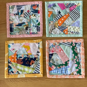 May include: Four handmade fabric coasters with patchwork designs. Each coaster features a unique arrangement of colourful fabric scraps, including floral, polka dot, and patterned textiles. The edges are finished with a contrasting border in pink, green, yellow, and peach.