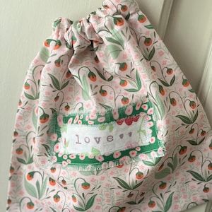 May include: A pink floral drawstring bag with green and orange accents. The bag has a patch with the word "love" embroidered on it. The bag is suspended against a white background.