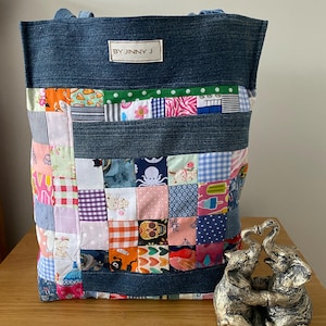 May include: A patchwork tote bag with a denim top and straps. The bag features a variety of colourful fabric squares with different patterns. The label on the bag reads "BY JINNY J". A silver elephant sculpture is next to the bag.