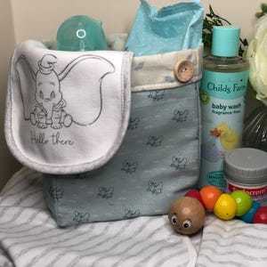 May include: A baby gift basket containing a Dumbo-themed bib, baby wash, and other baby essentials. The bib has a Dumbo illustration and the words "Hello there". The basket is light blue with a Dumbo pattern.