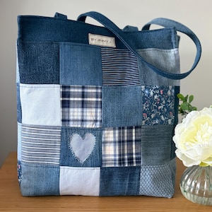May include: A patchwork denim tote bag with various blue fabric squares. The bag features a heart-shaped appliqué and handles. The label reads "BY JINNY J". The bag is sitting on a wooden surface.
