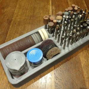May include: A gray plastic organizer filled with various rotary tool bits and accessories. The organizer contains cylindrical sanding drums, grinding stones, and other bits. The items are arranged on a wooden surface.