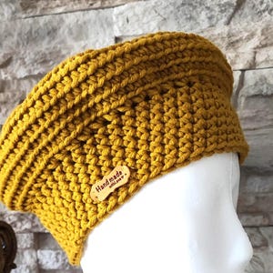 Puede incluir: Diadema de ganchillo de color amarillo mostaza. El calentador de oídos hecho a mano se muestra en una cabeza de maniquí blanca. Una pequeña etiqueta de madera con las palabras "Handmade with love" está adherida a la diadema. El fondo es una pared de piedra.