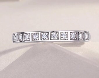 May include: A silver ring with a row of square-set, clear gemstones. The ring is set against a soft, neutral background, highlighting the sparkle of the stones. The design is simple and elegant.