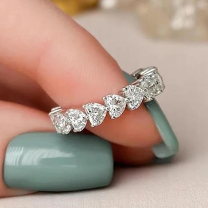 Może przedstawiać: Srebrny pierścionek z przezroczystymi kamieniami szlachetnymi w kształcie serca. Pierścionek jest trzymany na tle dłoni i jasnego zielonego lakieru do paznokci.
