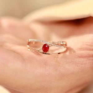 May include: A gold ring with a central red gemstone. The ring has a delicate, interwoven design, embellished with small, clear stones. The oval-shaped gemstone is a vivid red. The ring is shown against a light-toned backdrop.