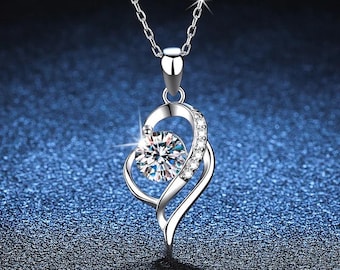 May include: A silver-colored pendant necklace featuring a heart-shaped design. The pendant has a large, clear, round gemstone at its center, surrounded by smaller, clear gemstones. The necklace chain is also silver-colored.