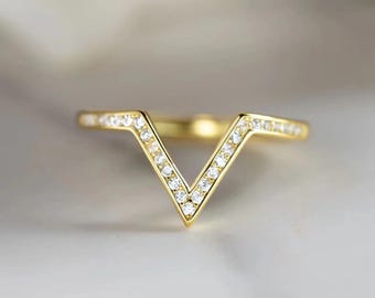 May include: A gold-colored ring with a V-shaped design. The ring is encrusted with small, clear gemstones along the V-shape and the band. The ring is set against a soft, blurred background.