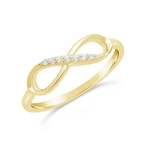 Puede incluir: Un anillo dorado con forma de símbolo de infinito. El anillo presenta pequeñas piedras preciosas transparentes engastadas a lo largo de la curva superior del diseño infinito. El anillo está sobre un fondo blanco.
