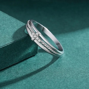 Puede incluir: Un anillo plateado con un diseño de cruz. El anillo presenta una doble banda, una con pequeñas cuentas y la otra con piedras transparentes que forman una cruz. El anillo se exhibe sobre una superficie verde azulado.