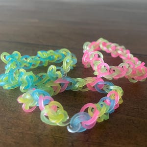May include: Three colorful loom band bracelets. One bracelet is a mix of blue, green, and yellow. Another is pink and green. The third bracelet is a mix of pink, blue, and yellow. The bracelets are on a brown wooden surface.