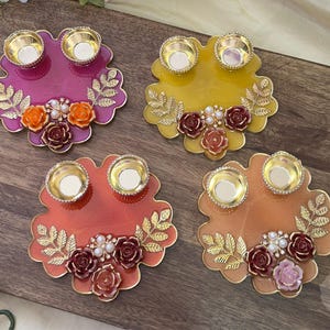Op de afbeelding: Vier decoratieve kandelaars in roze, geel, oranje en perzik. Elke houder heeft twee goudkleurige kaarsbekers, bloemaccenten en gouden blad details. De houders zijn gerangschikt op een houten oppervlak.