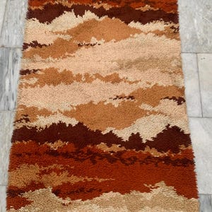 May include: A rectangular, textured rug with a landscape design in shades of brown, beige, and orange. The rug has a shaggy appearance and is likely made of a soft material. The color palette evokes a warm, earthy feel.