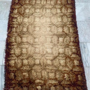 May include: A rectangular shag rug in shades of brown and tan. The rug has a textured, plush surface with a darker brown border and a repeating geometric pattern in the center. The rug's design creates a warm and inviting aesthetic.