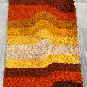May include: A rectangular shag rug featuring a horizontal wave pattern in shades of brown, orange, yellow, and cream. The high-pile texture creates a dense, tufted surface with a repeating gradient of warm earth tones across the entire piece.