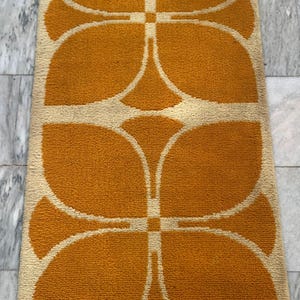 May include: A rectangular rug with a repeating floral pattern in shades of orange and cream. The design features four large, stylized flower shapes, creating a symmetrical and eye-catching visual. The rug's texture appears soft and plush.