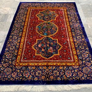 Könnte beinhalten: Ein rechteckiger Teppich mit einem tiefroten Mittelfeld und drei dunkelblauen Medaillons, die komplexe Blumenmuster enthalten. Der Teppich hat einen breiten dunkelblauen Rand mit bunten Blumenmotiven und weiße Fransen an beiden Enden.