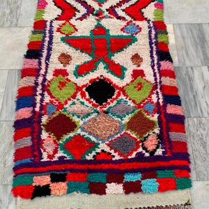 May include: A vibrant, rectangular rug with a central design featuring a red star and geometric shapes. The rug showcases a palette of red, green, blue, and purple, with a cream-colored background and fringed edges. The rug's dimensions are not visible.