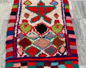 May include: A vibrant, rectangular rug with a central design featuring a red star and geometric shapes. The rug showcases a palette of red, green, blue, and purple, with a cream-colored background and fringed edges. The rug's dimensions are not visible.