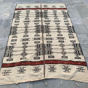 Peut inclure: Un tapis rectangulaire crème avec des motifs géométriques noirs, marron et rouges. Le tapis présente un motif central avec des formes de losanges et rectangulaires, bordé de bandes de couleur et de motifs décoratifs. Des glands sont visibles au bord inférieur.